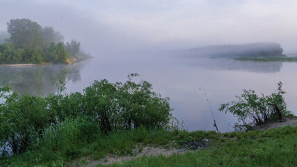 A beautiful, misty river at dawn. Lush green vegetation and a fishing rod are visible on the quiet, peaceful banks © Александр Арендарь