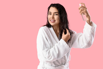 Young woman in bathrobe applying hair spray on pink background