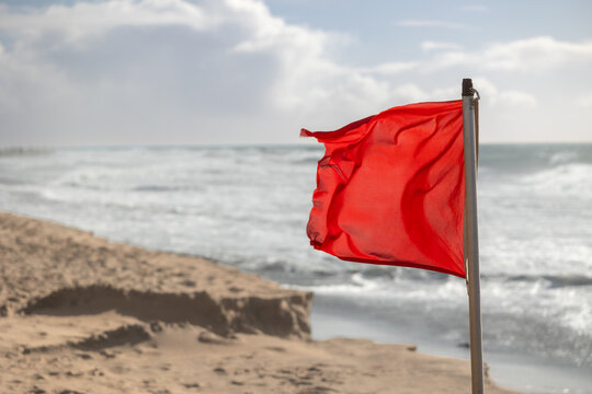 Red flag prohibiting swimming on sandy beach against stormy sea on sunny day. No people