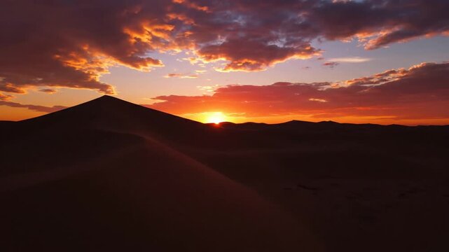 Bright golden hour sunset over rolling sand dunes in a vast desert landscape with blowing sand on ridges, scattered clouds in an orange sky, and deep shadows at dusk.