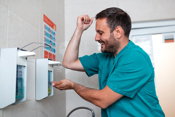 Male surgeon or nurse in green scrubs washing hands with professional disinfectant dispenser in a...