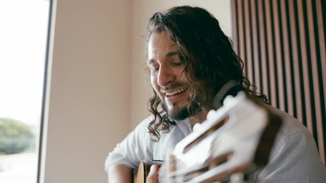 Talented young male artist with long curly hair playing an acoustic guitar and singing passionately. Music composer creating a new song at home