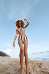 Naklejka premium Young woman in striped bikini stands on sandy beach with raised arm smiling happily woman enjoying summer vacation near calm sea water under blue sky with clouds.