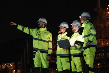 Four  workers in bright yellow coveralls and white helmets inspect industrial refinery site at...