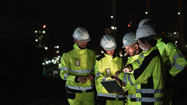 Five Construction Workers Examine Construction Site with Laptop, Reviewing Plans in Dimly Lit Industrial Area, Wearing Hard Hats and Yellow Vests, Conducting Inspection Work at Nighttime.