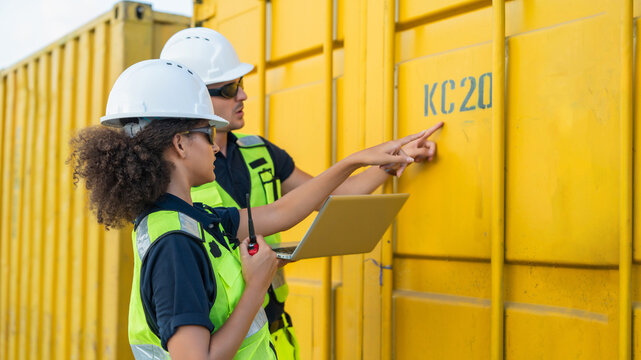Woman and Man Inspecting Yellow Container with "KC20 1" While Pointing at Laptop and Using Radio for Cargo Logistics Monitoring Near Shipping Container in Port Environment. construction worker wearing