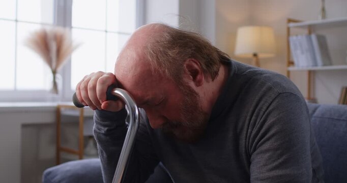 Sad distressed old senior man at home. Older man on sofa leaning on cane takes off glasses suffering, feeling lonely, left and forgotten by family, thinking of illness, or grieving for deceased wife