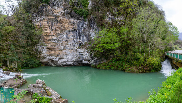 Scenic view of a small turquoise karst lake at the foot of a high limestone cliff. Blue lake water surrounded by lush spring greenery and rocks. Bzyb River area, Abkhazia.