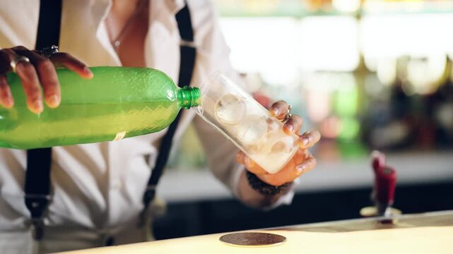 Professional bartender pouring a sparkling green beverage from a plastic bottle into a tall glass filled with ice cubes on a bar counter