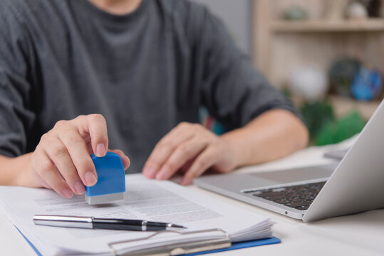 Close up hand of startup founder stamping business report with laptop on table. Male freelancer working on administrative document in casual t-shirt. SME deal confirmation.