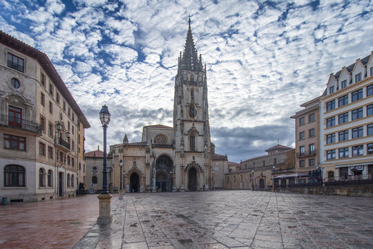 Catedral de San Salvador de Oviedo en la Plaza de Alfonso II el Casto, Asturias, destacando su torre g&oacute;tica, fachadas hist&oacute;ricas y el suelo de piedra mojado bajo un cielo de nubes algodonosas.