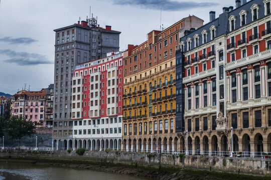Fachadas de edificios tradicionales y modernos en la Ribera del Nervi&oacute;n, Bilbao, mostrando sus caracter&iacute;sticos colores, balconadas y soportales junto al cauce de la r&iacute;a bajo un cielo nublado.