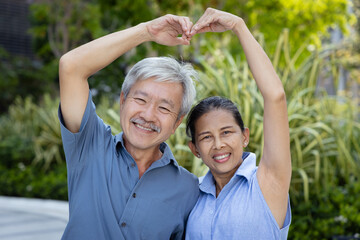 Obraz premium Happy and lovely married Asian senior couple, old casual husband and wife in urban park with good quality of life, making Asian style big heart body gesture.