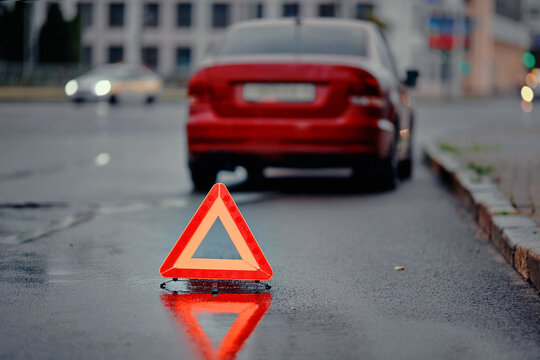 Reflective warning triangle behind broken vehicle. Car accident aftermath on dark, wet city road with emergency triangle marking hazard zone, ensuring visibility for oncoming drivers. Selective focus