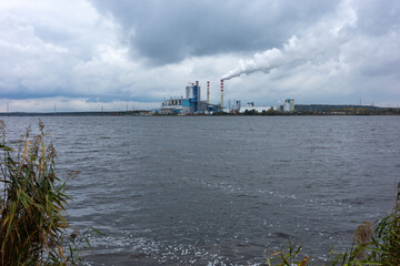 Obraz premium A view of a distant coal-fired power plant located on the shore of a lake. Exhaust fumes from a smoking chimney stand out against a cloudy sky.