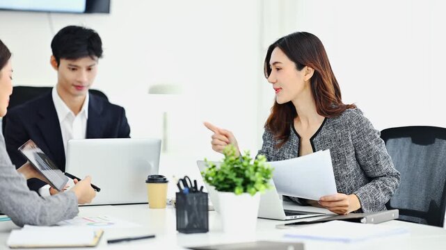 Asian business team discussing documents during a meeting in modern office. Businessman explaining paperwork to female colleague while working together with laptop and financial reports. Teamwork, cor