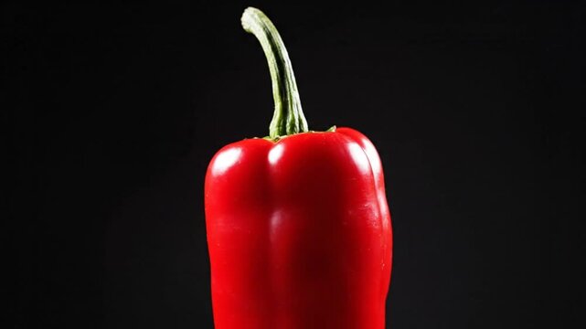 Red bell pepper with a curved green stem illuminated against a black background with shallow depth of field.