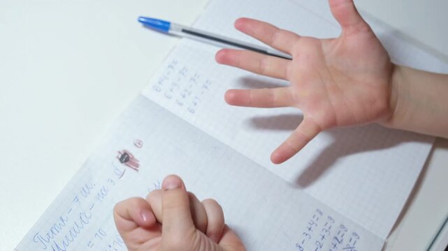 Close-up of a child counting on fingers while solving basic arithmetic homework, hands over a notebook with simple math exercises