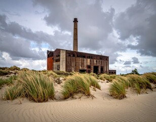 Fototapeta premium Old factory stands in sand with tall grass under cloudy sky near coast during afternoon light