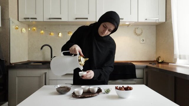 muslim woman in black hijab pouring arabic coffee from white dallah into a small finjan cup on the kitchen table during traditional ramadan serving