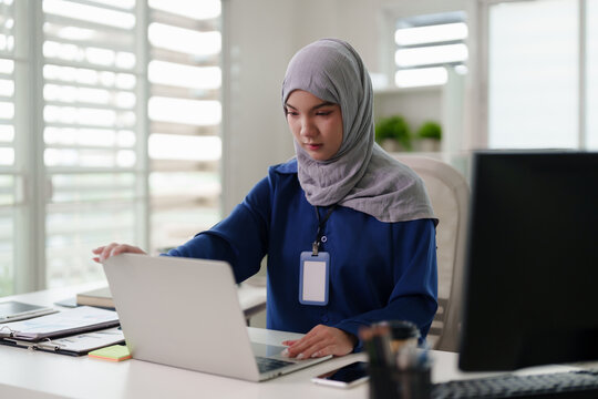 Muslim adult woman professional working on laptop at modern office desk Asian employee managing business projects online creating reports planning strategy