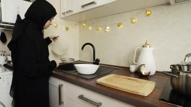muslim woman in black hijab rinses rice through a strainer at the sink while warm ramadan-style decor glows in the background, showing a calm home cooking routine