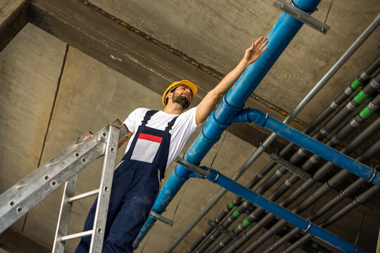 Construction worker on a ladder inspecting ceiling pipes for quality control.