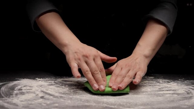 Closeup of baker hands shaping vibrant green dough on floured dark countertop, capturing the careful kneading process in a moody, softly lit kitchen scene