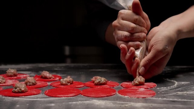 Close-up of chef hands piping meat filling onto bright red dough circles on floured surface, preparing handmade stuffed dumplings or ravioli in dark kitchen