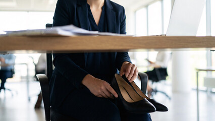 Fototapeta premium Young Professional Woman Removing High Heels Under Desk In Modern Office, Workplace Discomfort, Foot Pain Relief, Corporate Environment, Office Lifestyle, Candid Work Moment