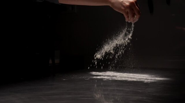 Slow motion chef hand sprinkling fine white flour across dark kitchen countertop, creating a dramatic cloud of powder in low key cooking preparation scene