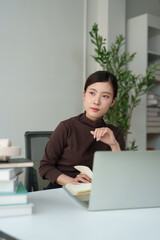Fototapeta premium Young Asian woman student learning and thinking while studying with a laptop and books at a bright desk, focusing on online education and planning future career goals at home