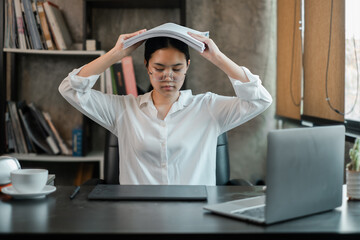 Obraz premium A young woman in an office setting appears stressed, holding documents over her head, surrounded by a laptop and books.