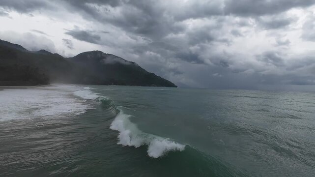 Storm waves approaching tropical coastline. Aerial drone view of rolling ocean swell under heavy clouds with mountain backdrop, creating dramatic seascape with rough water and storm atmosphere.