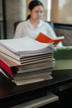 A professional woman reading documents at her desk, surrounded by a stack of files in a modern office setting.