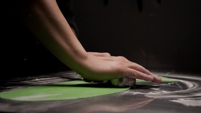 Close-up of a baker hands cutting shapes from rolled green dough on a floured work surface in a dimly lit kitchen, highlighting texture and craftsmanship