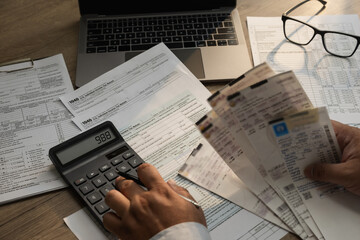 Obraz premium Close-up of a person calculating expenses with a calculator while holding multiple receipts and reviewing tax documents on a desk with a laptop, suitable for expense reporting, budgeting, reimbursemen