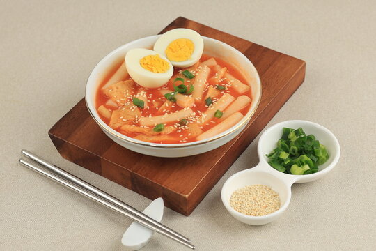 A bowl of toppoki served with a boiled egg, sprinkled with sesame seeds and sliced ​​spring onions, on a wooden cutting board. Isolated on a clean light gray background