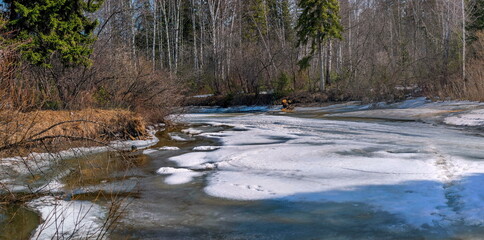 Fototapeta premium a frozen pond with patches of snow and ice. Trees and shrubs grow all around. The shadows of the trees are visible.