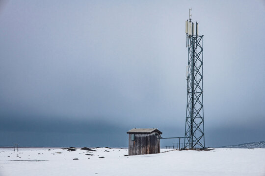 Abandoned utility tower and small wood hut in a snowy Icelandic countryside landscape along the Ring Road, minimalist winter scene with frozen ground, overcast sky, rural isolation, remote Nordic mood