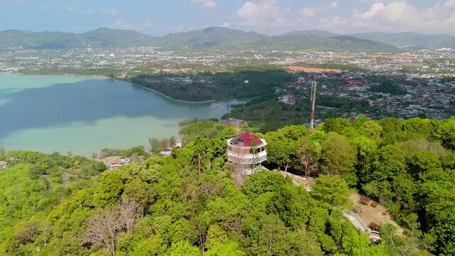 Panoramic aerial Kaokard Viewpoint Tower, a stunning vista unfolds over Phuket city, a tranquil bay, distant mountains. Lush green forest envelops the tower under bright blue sky filled white clouds.