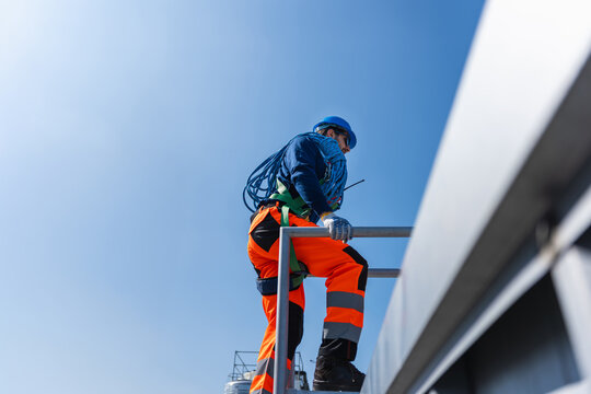 Industrial worker in safety harness and helmet carrying blue rope on a high platform, Professional technician in reflective orange uniform climbing industrial structure for maintenance