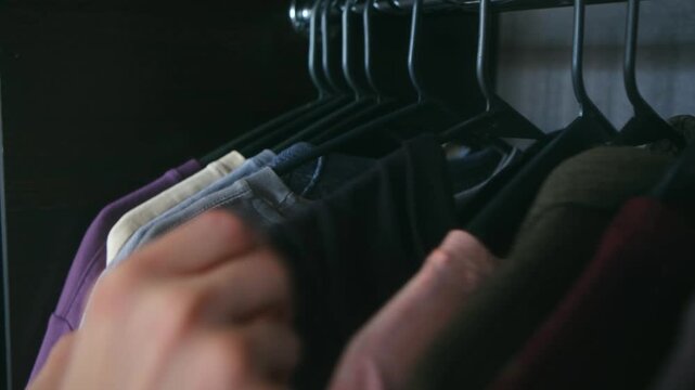 Close up person hand browsing through row of colorful shirts on hangers inside dark closet, finally selecting one and taking it off rail to get dressed for day. Person choosing clothes from wardrobe