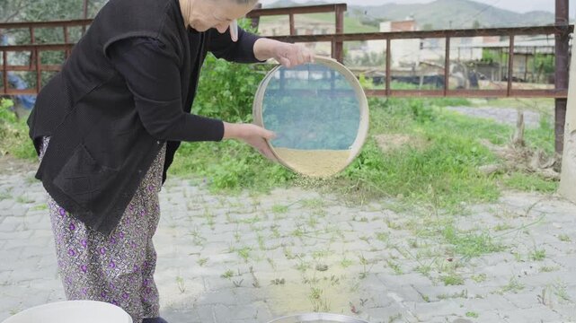 Old woman sifting wheat with a traditional sieve in her hands in a rural village environment, representing the timeless connection between human labor and sustainable farming