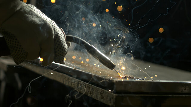 A person uses a welding torch in a workshop. Sparks and smoke rise as metal is fused together. The background shows blurred lights and shadows, creating a focused work environment.