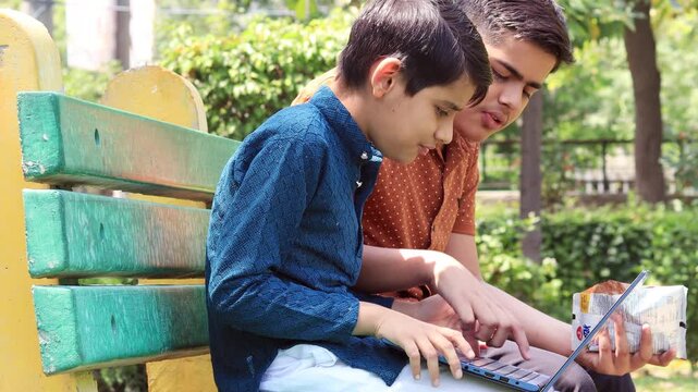 Two elementary age child of Indian ethnicity sitting outdoor in nature and using laptop together and eating snacks.