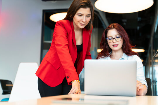 A woman in a red blazer assists another woman with glasses at a laptop on a conference table in a modern office. They are coworkers engaged in a work project.