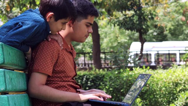 Two elementary age child of Indian ethnicity sitting outdoor in nature and using laptop together video portrait.