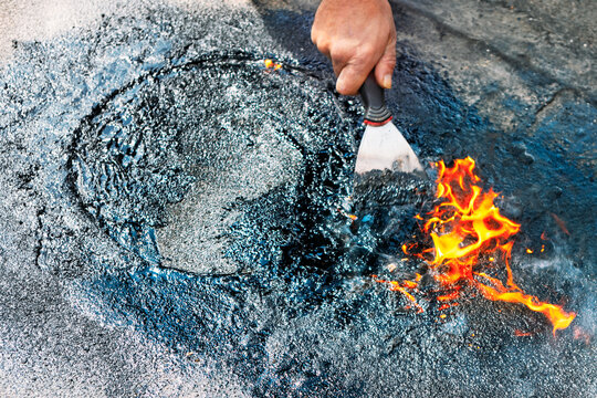 Worker spreads hot tar with a metal spatula as bright flames lick the surface of the asphalt. Molten material glows against the rough, gray texture of the road