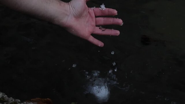 Close up of a hand releasing a small snowball into water, capturing the moment it slips away and begins to melt. Gentle ripples and dark tones create a calm, atmospheric scene.